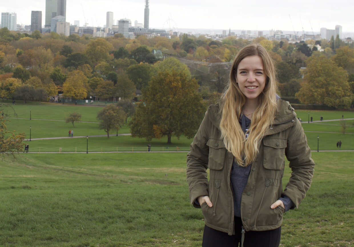 Photo of the Beyond Page One author, Kaitlyn Nightingale. She is wearing a green jacket and is standing infant a grassy park with trees and skyscrapers in the background.